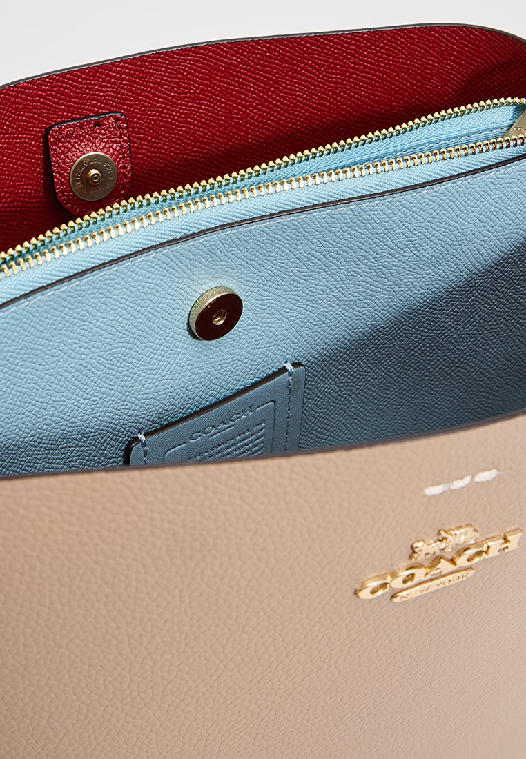 Sac à main en cuir beige de Coach avec une poche zippée à l'intérieur bleu et une doublure rouge, mettant en avant un logo doré et une fermeture à bouton magnétique.