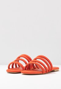 Sandali in suede arancione con quattro ampie cinghie imbottite e suole piatte, caratterizzati da una texture liscia e un design minimalista.