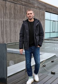 Černá prošívaná bunda s kapucí, černý svetr, modré džíny a bílé tenisky se zelenými akcenty. Venkovní prostředí s dřevěným pozadím.