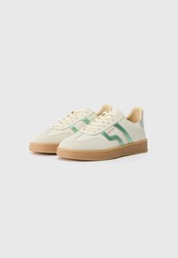 Kremfargede sneakers med grønne striper, en tan gummisåle og detaljer i metallisk sølv på hælen. Snøring med en glatt tekstur.