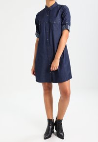 Vestido camiseros de mezclilla azul oscuro con mangas cortas, frente de botones, dos bolsillos en el pecho y puños enrollados. Llevado con botas negras hasta el tobillo.