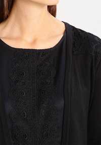 Svart cardigan med texturerad spetsdetalj på axlarna och en dekorativ broderad panel fram. Slät tygfinish.