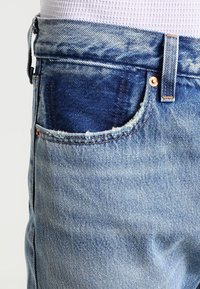 Close-up of light blue denim jeans with a frayed front pocket edge and copper rivets, worn with a white textured top.