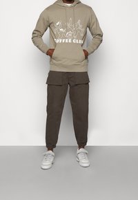 Khaki bluza z kapturem z białym nadrukiem "COFFEE CLUB". W zestawie ciemne spodnie typu cargo z wieloma naszywanymi kieszeniami i elastycznymi mankietami. Białe trampki.