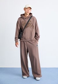 Brun hættebluse med grafisk tryk, parret med matchende vidde sweatpants. Færdig med en sort crossbody taske og lyse sko.