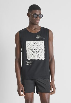 Muškarac koji nosi crnu majicu bez rukava s bijelim uzorkom kukičanog kvadrata, crne kratke hlače i tamne sunčane naočale, stoji ispred jednostavne pozadine.