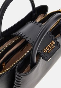 Černá kožená kabelka se zlatým zipem a prošívaným klopou, na které je zlato znak "GUESS EST 1981", prohlédnuta z horního úhlu.
