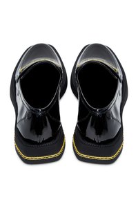 Chaussures en cuir verni noir à bout ouvert, avec des coutures jaunes sur une semelle noire épaisse. Texture lisse et forme arrondie.
