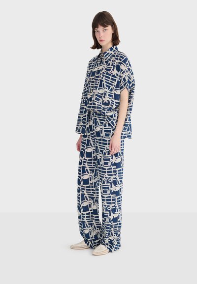 Kobieta stojąca, ubrana w luźną koszulę w granatowo-biały wzór oraz dopasowane szerokie spodnie, do tego białe plecione buty.