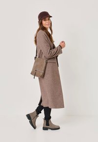 Palton din lână maro, pantaloni negri ajustați, cizme gri până la glezne și o șapcă maro închis. Îmbrăcămintea include o geantă gri tip crossbody cu fermoar.