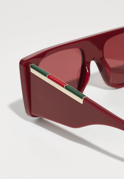 Ochelari de soare roșii cu lentile dreptunghiulare. Rama din plastic gros are un accent aurit cu dungi verzi și roșii pe brațele ochelarilor.