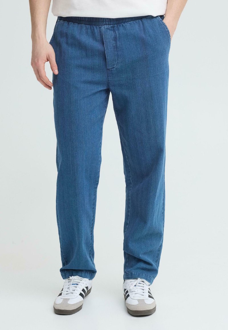 Mann iført blå denimbukser med elastisk midje, sidelommer og hvite joggesko med svarte striper, står mot en ensfarget bakgrunn.