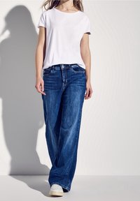 Weißes T-Shirt mit kurzen Ärmeln, kombiniert mit weiten, ausgebleichten blauen Jeans. Das Outfit zeichnet sich durch ein klassisches Design und eine lässige Passform aus, abgerundet durch weiße Turnschuhe.