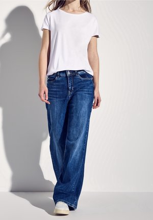 Camiseta blanca de manga corta combinada con jeans de pierna ancha y de color azul desgastado. El conjunto presenta un diseño clásico y un ajuste informal, complementado con zapatillas blancas.