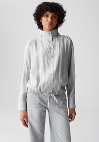 Hellgraue Bluse mit Knöpfen, hochgeschlossenem Kragen, elastischem Taillenbund, langen Ärmeln und weicher, glatter Textur. Die Vorderseite verfügt über mehrere Knöpfe.