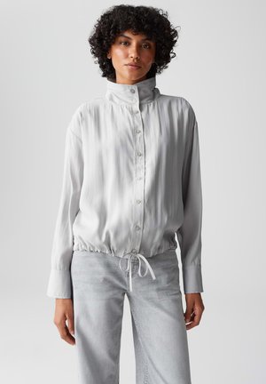Hellgraue Bluse mit Knöpfen, hochgeschlossenem Kragen, elastischem Taillenbund, langen Ärmeln und weicher, glatter Textur. Die Vorderseite verfügt über mehrere Knöpfe.