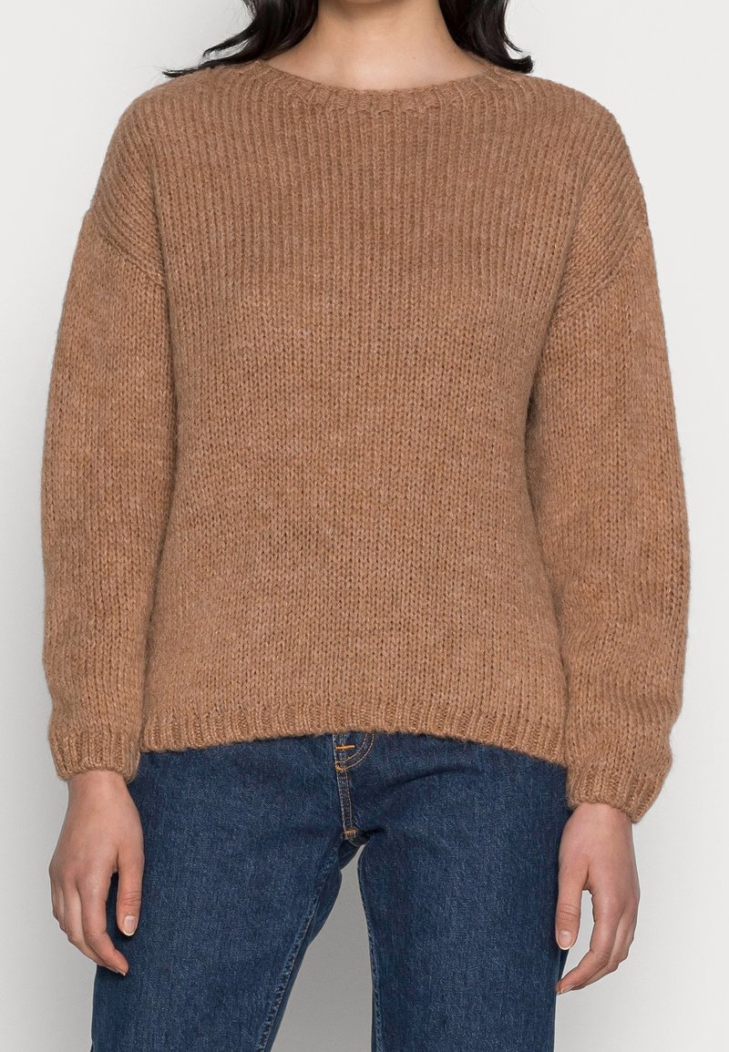 Femme portant un pull marron en maille à manches longues associé à un jean bleu, debout avec les bras détendus le long du corps.