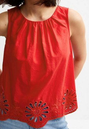 Czerwony top bez rękawów z marszczonym dekoltem i kwiatową haftowaną ażurową wstawką na falowanym brzegu, noszony z niebieskimi jeansami.
