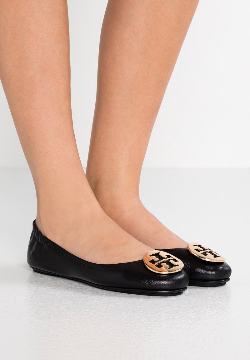 Pieds chaussés de ballerines en cuir noir avec des logos décoratifs ronds dorés sur les orteils, posés sur une surface blanche.