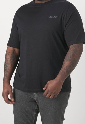 T-shirt noir à manches courtes Calvin Klein porté par une personne avec des bras tatoués, associé à un jean gris foncé sur un fond uni.