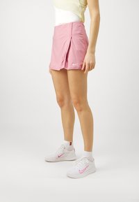 Rosa Sportshorts mit einem gefältelten Design, versehen mit einem kleinen weißen Nike-Logo. Kombiniert mit weißen Turnschuhen mit pinken Akzenten.