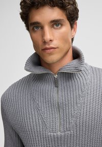 Pullover grigio a maglia con colletto alto e chiusura con zip. Presenta costine testurizzate e un motivo a V sulla parte frontale.