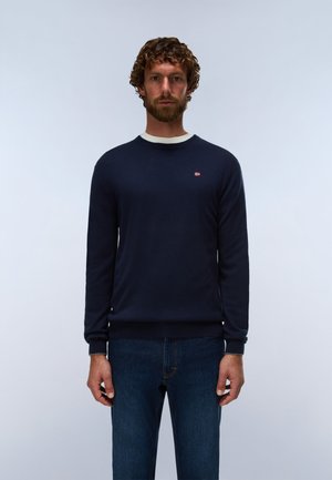Man met krullend haar en baard, die een donkerblauwe trui draagt met een klein rood-wit logo op de borst en een blauwe spijkerbroek tegen een effen achtergrond.