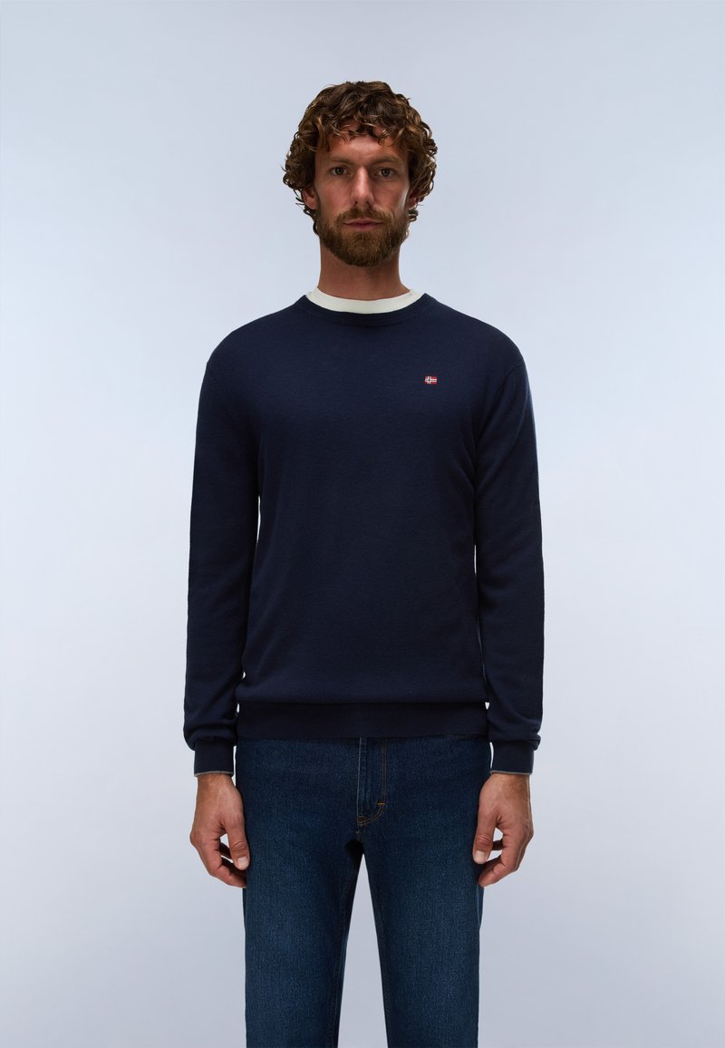 Mann mit lockigem Haar und Bart, der einen dunkelblauen Pullover mit kleinem rot-weißem Logo auf der Brust und blaue Jeans vor einem einfarbigen Hintergrund trägt.