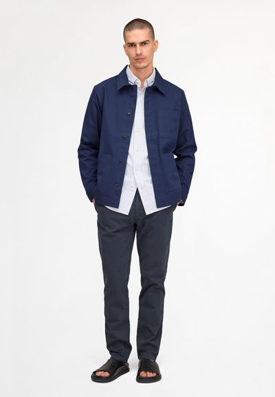 Uomo in piedi con una giacca blu navy sopra una camicia bianca a righe, pantaloni scuri e sandali neri aperti sulle dita, su sfondo bianco.