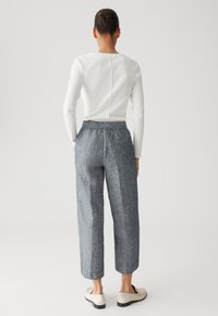 Personne de dos, portant un haut blanc à manches longues, un pantalon large gris et des mocassins blancs sur un fond uni.