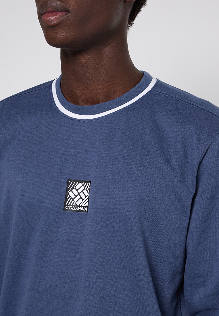 Persona che indossa una felpa blu con collo a girocollo, con un colletto a strisce bianche e un piccolo logo Columbia in bianco e nero centrato sul petto.