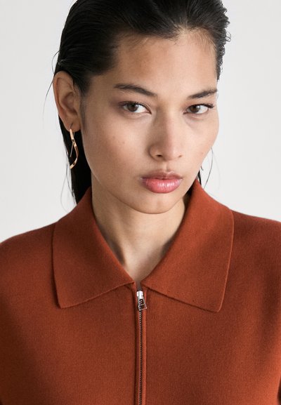 Jeune femme aux cheveux mouillés portant des boucles d'oreilles en or en forme de vagues et un pull zippé à col de couleur rouille, devant un fond uni.
