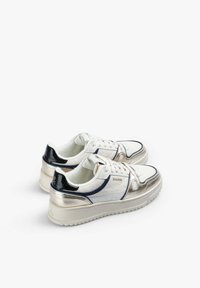 Zapatillas de deporte bajas con tela blanca, acentos metálicos dorados, detalle del talón en negro, suela beige y cordones blancos, dispuestas lado a lado sobre un fondo blanco.