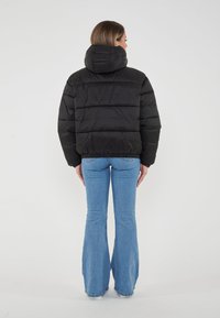 Giacca a vento nera con cappuccio, design corto e imbottitura orizzontale. Indossata con jeans chiari a zampa d'elefante, vista da dietro.