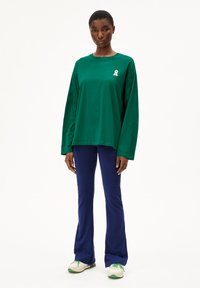 Groene langemouwenshirt met een wit lintlogo, gecombineerd met marineblauwe flared broek en witte sneakers met groene accenten.