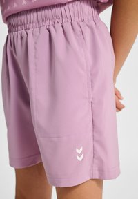 Lichtpaarse shorts gemaakt van gladde stof, met een elastische tailleband en een wit chevron-logo aan de zijkant. Eenvoudig, functioneel ontwerp.