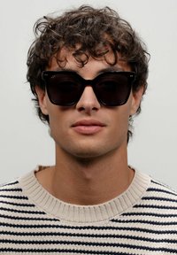 Černé oversize sluneční brýle s geometrickým rámem. Modelka má na sobě béžový pruhovaný svetr. Krátké kudrnaté vlasy a neutrální výraz.