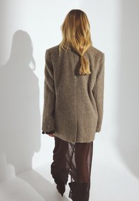 Femme aux longs cheveux blonds attachés en queue de cheval basse, portant un manteau marron texturé et un pantalon noir transparent, debout contre un mur blanc avec une ombre.