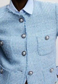 Veste en tweed bleu clair texturée avec boutons couleur perle, poche avant et col. Présente un ourlet effiloché et une coupe ajustée.