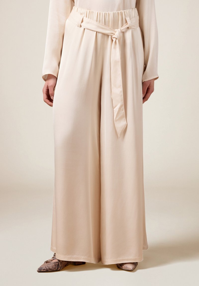 Brede broek in een zachte, lichtbeige stof met een elastische tailleband en riemdetail. Glad textuur met een ontspannen pasvorm.