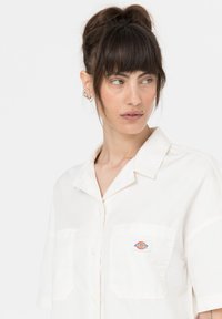 Camicia a maniche corte color crema con bottoni, dotata di due tasche frontali e un piccolo logo colorato sulla tasca sinistra. Tessuto morbido e liscio.