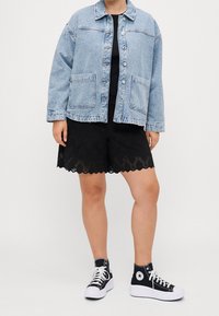 Lichtblauwe spijkerjack met knoopsluiting, twee steekzakken aan de voorkant, gedragen over een zwart shirt en zwarte kanten shorts, gecombineerd met zwarte hoge sneakers.