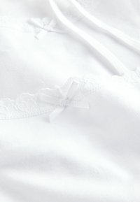 Witte camisole met kantafwerking en een klein strikdetail. De stof is glad met een lichte glans en heeft verstelbare spaghettibandjes voor een goede pasvorm.