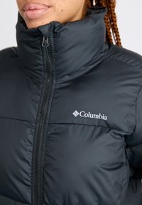 Giacca a puffo nera con colletto alto, chiusura con zip e logo Columbia sul lato sinistro del petto. Presenta un tessuto in nylon texturizzato.