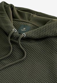 Mørkegrøn quiltet hættejakke med snore, som har et mærke mærket "NEXT." Stoffet har en tekstureret overflade med et blødt, fleksibelt design.