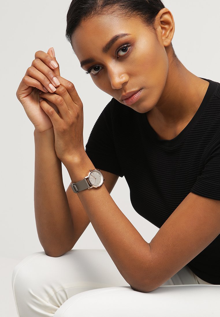 Silver round watch with a minimalist face, gray leather strap, and rose gold accents, worn on a wrist against a neutral background.