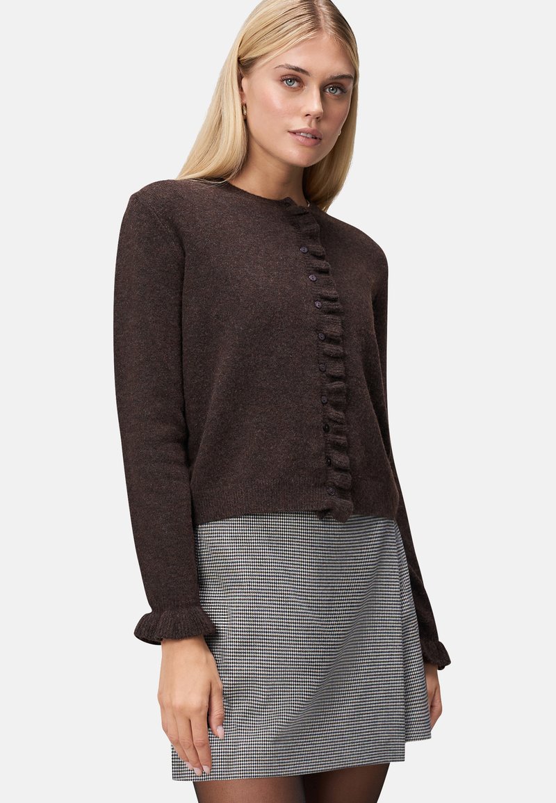 Cardigan lavorato a maglia marrone con scollo rotondo, chiusura a bottoni e dettagli arricciati lungo la parte anteriore. Indossato sopra una gonna a scacchi bianca e nera.