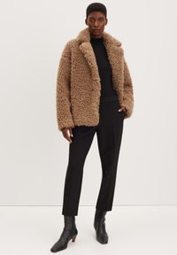 Cappotto marrone peloso con un ampio colletto, abbinato a un dolcevita nero, pantaloni neri e stivali neri alla caviglia. Design strutturato e oversize.