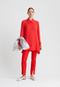 Rotes Langarmhemd mit spitzem Kragen, kombiniert mit passenden Slim-Fit-Hosen mit Seitenschlitzen. Silberne flache Schuhe, dazu eine metallische Tasche.