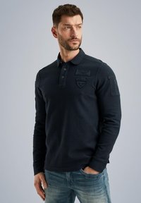Navyblaues Langarm-Polo aus strukturiertem Stoff, mit einem Logo auf der Brust und zwei Knöpfen. Kombiniert mit hellblauen Jeans.